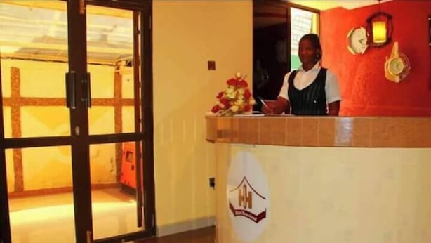 Recepção de hotel com divisória de vidro, recepcionista e parede vermelha com relógios.