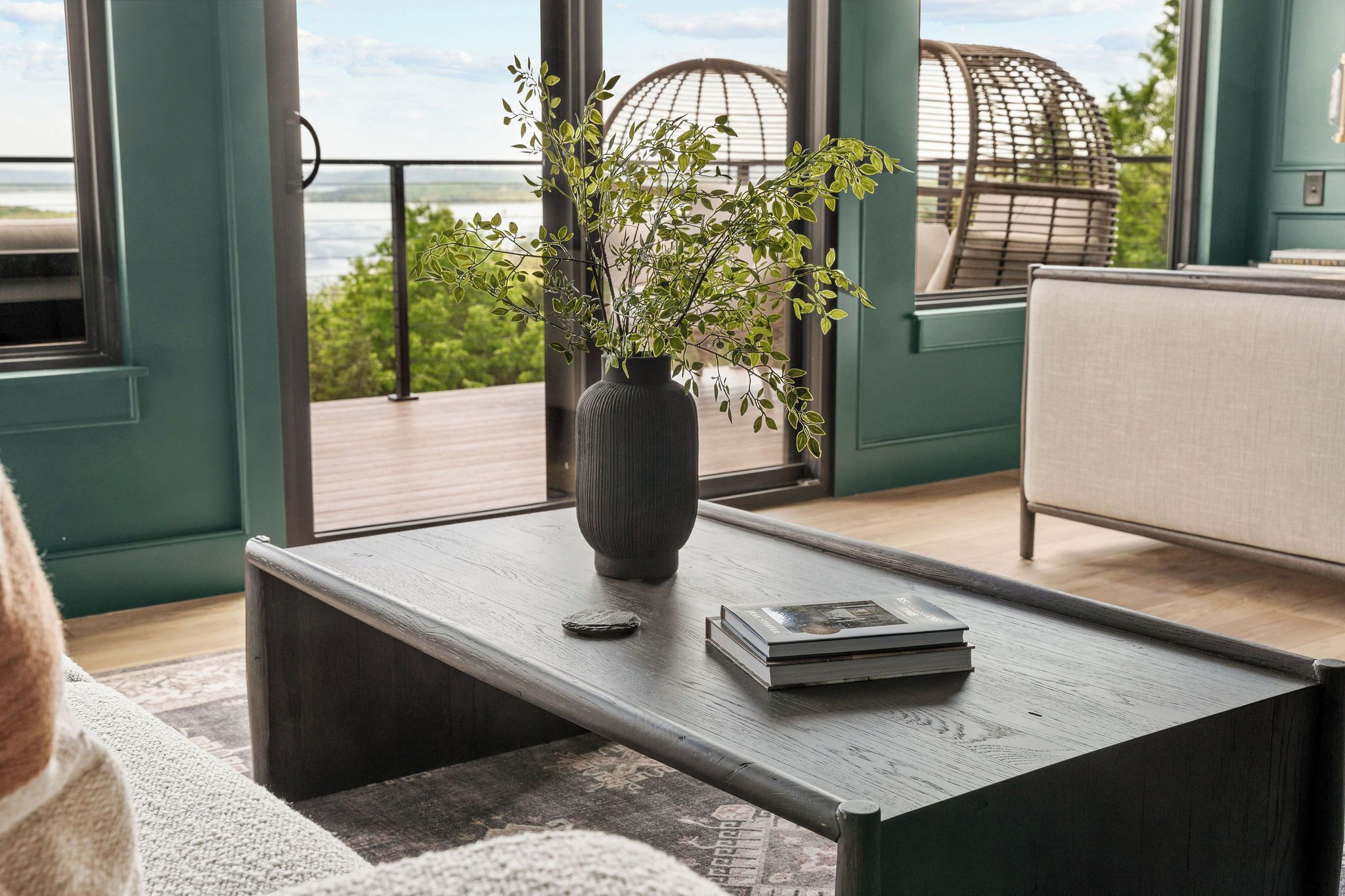 Un salon moderne doté d’une grande fenêtre, d’une table basse en bois foncé, d’un vase orné de feuillage vert et offrant une vue sur l’extérieur.