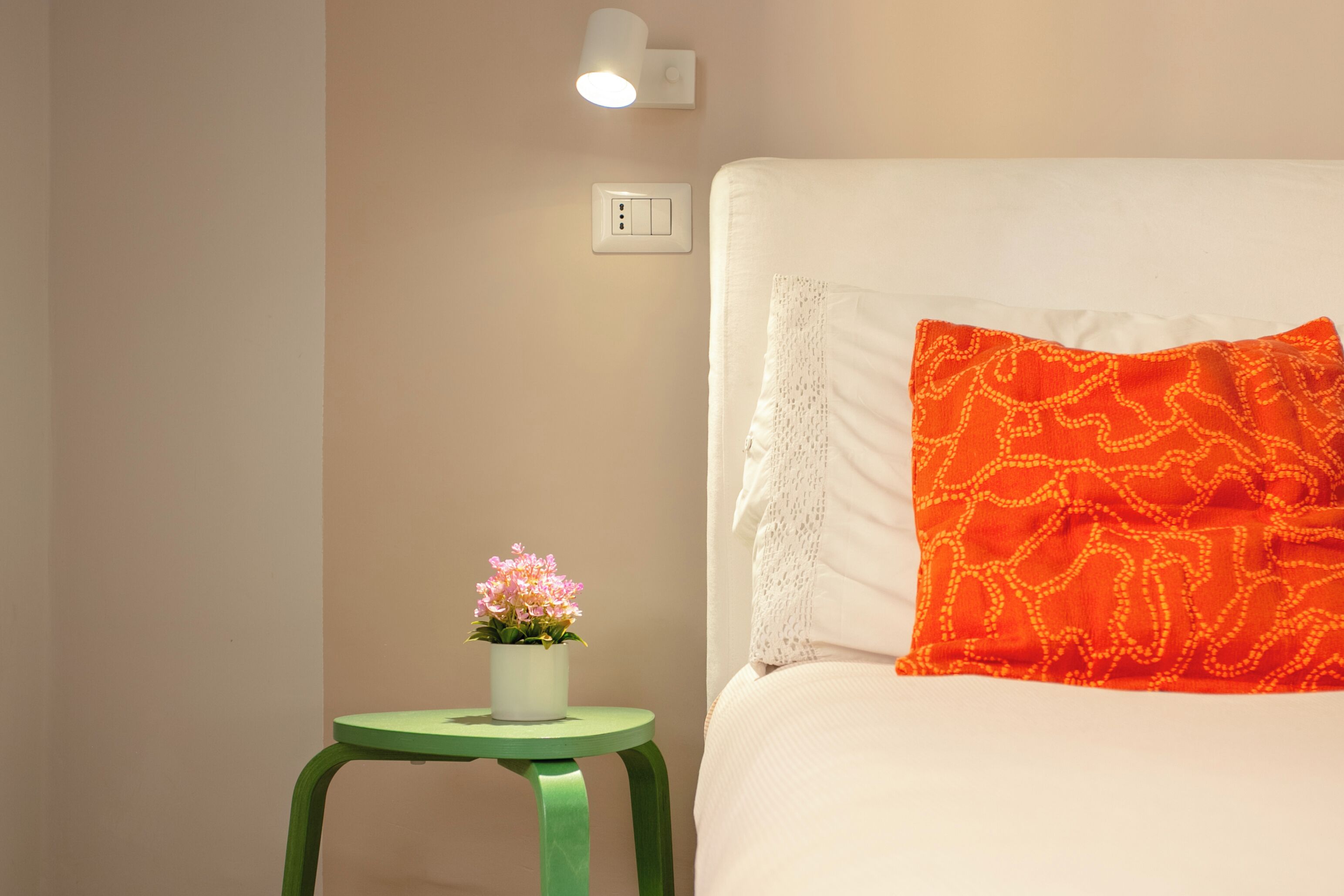 Una cama bien hecha con un cojín blanco y naranja, un taburete verde pequeño con una planta en maceta, y una lámpara fijada a la pared.