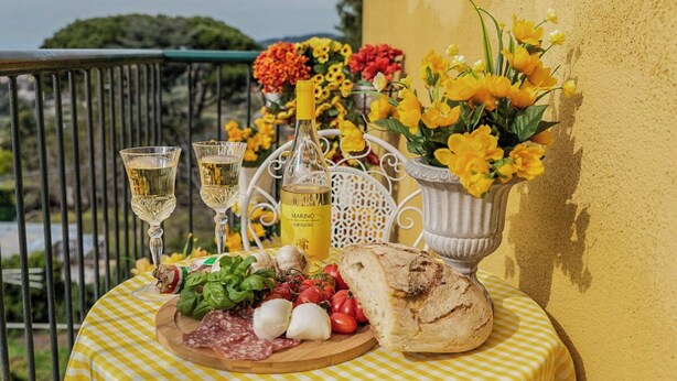 A table with a checkered tablecloth, a bottle of olive oil, a bouquet of yellow flowers, a loaf of bread, and a charcuterie board with cheese, tomatoes, and basil. - Casa Marino (Ciampino)