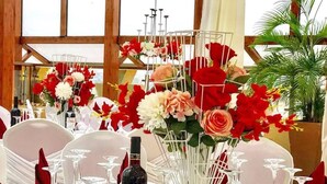 Una mesa de comedor dispuesta con platos blancos, servilletas rojas y copas de vino, rodeada de centros de mesa florales.