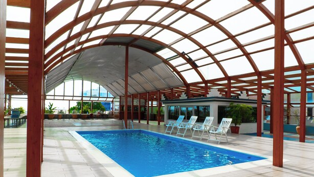 Una piscina interior con techo de vidrio, rodeada de sillas blancas y plantas en macetas.