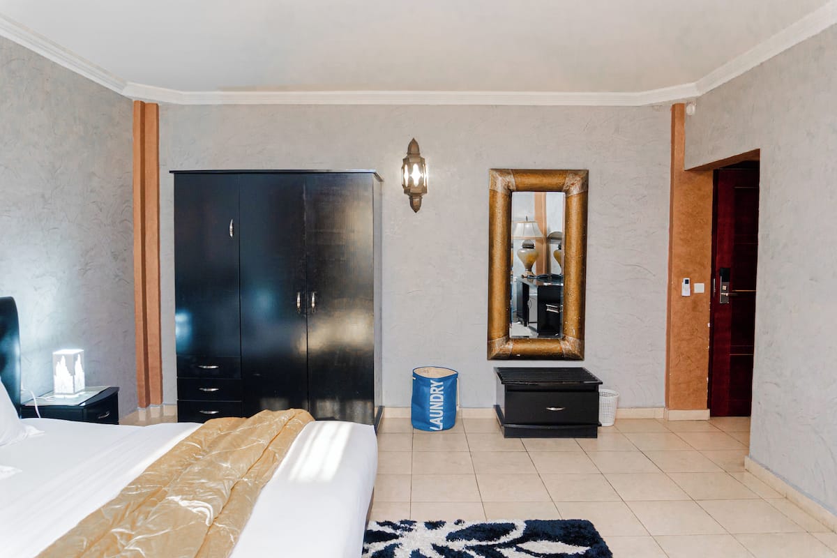 A hotel room with a bed, a wardrobe, a mirror, a laundry basket, and a small table with a lamp.