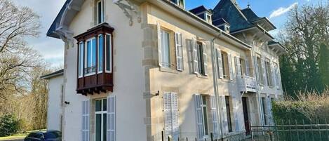 A large, multi-story house with a symmetrical facade, multiple windows with shutters, and a prominent balcony.