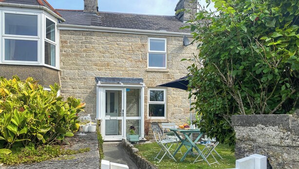 Une petite maison en pierre avec une porte d’entrée en verre, un petit patio avec une table et des chaises, et une enseigne indiquant « GRACE COTTAGE ».