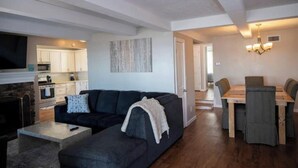 A modern living room with a dark sofa, a coffee table, and a fireplace. There is a dining area with a wooden table and chairs. The kitchen is visible in the background with white cabinets and a stone fireplace. - Spacious Vermilion House on the Lake (Vermilion)