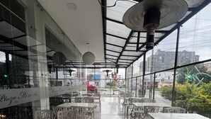 Een ruime eethoek buiten met witte tafels en stoelen, grote ramen met uitzicht op de stad, en een overdekt gedeelte met een plafondventilator.
