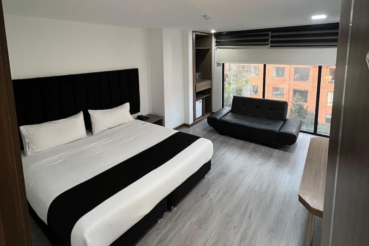 Ein modernes Schlafzimmer mit einem großen Bett, einer Couch und Blick auf Gebäude durch große Fenster.