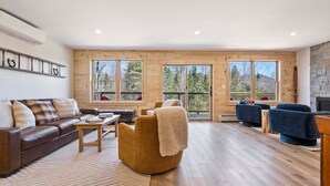 A spacious living room with a brown leather sofa, a round ottoman, and blue armchairs. Large windows offer a view of trees and a balcony. - Pointers Den-Fireplace, Arcade, 2 Primary Suites (Bethel)
