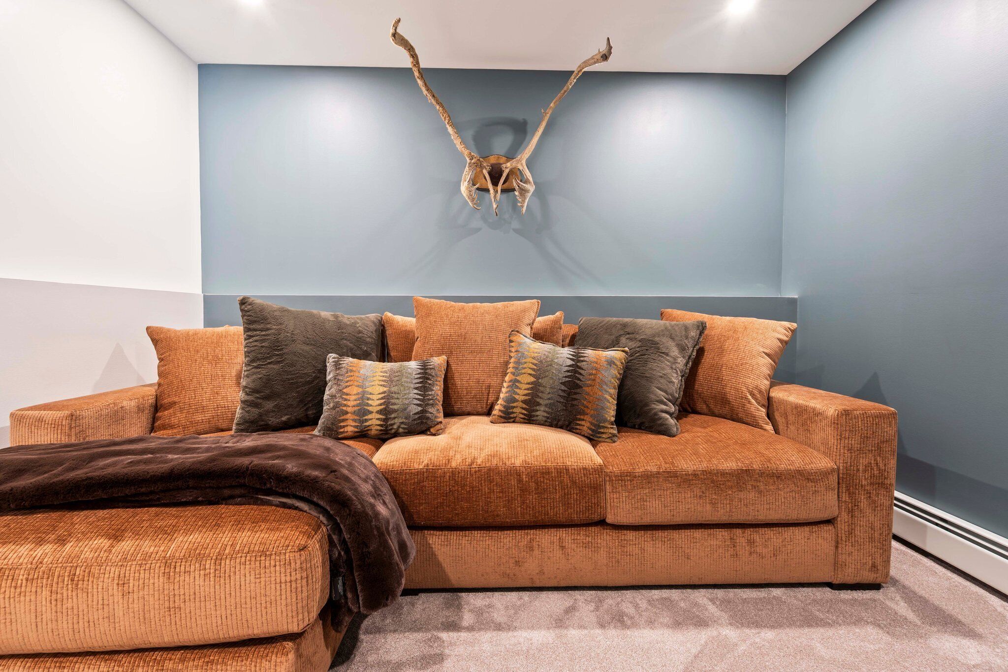 A modern living room with a large, orange sofa, multiple cushions, and a decorative antler wall piece.