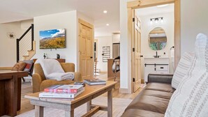 A cozy living room with a wooden coffee table, a brown sofa, and a white armchair. A staircase is visible in the background, leading to another room. - Pointers Den-Fireplace, Arcade, 2 Primary Suites (Bethel)