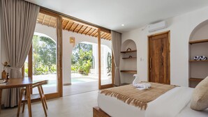 A bedroom with a bed, a wooden desk, and a view of a garden through a sliding door. - Luxurious 2BR Villa - Scenic Rice Field Views - Beautiful Swimming Pool & Garden (Canggu)