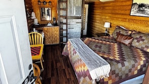 A cozy cabin interior with a wooden ceiling, a bed with a patterned blanket, a dining area with a table and chairs, and a kitchen area with a stove and a ladder.