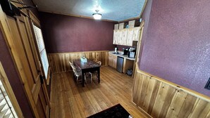 Une pièce comprenant une table à manger en bois, des chaises, un coin cuisine avec des placards et une fenêtre équipée de stores.