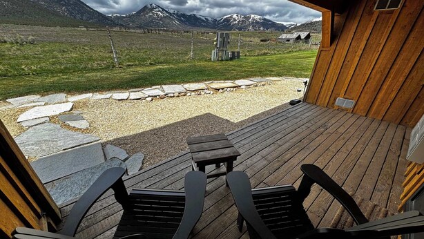 Une terrasse en bois avec deux chaises noires et une table, donnant sur un paysage montagneux agrémenté d’un sentier en pierre et d’une clôture.