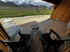 A wooden deck with two black chairs and a table, overlooking a mountainous landscape with a stone pathway and a fence.