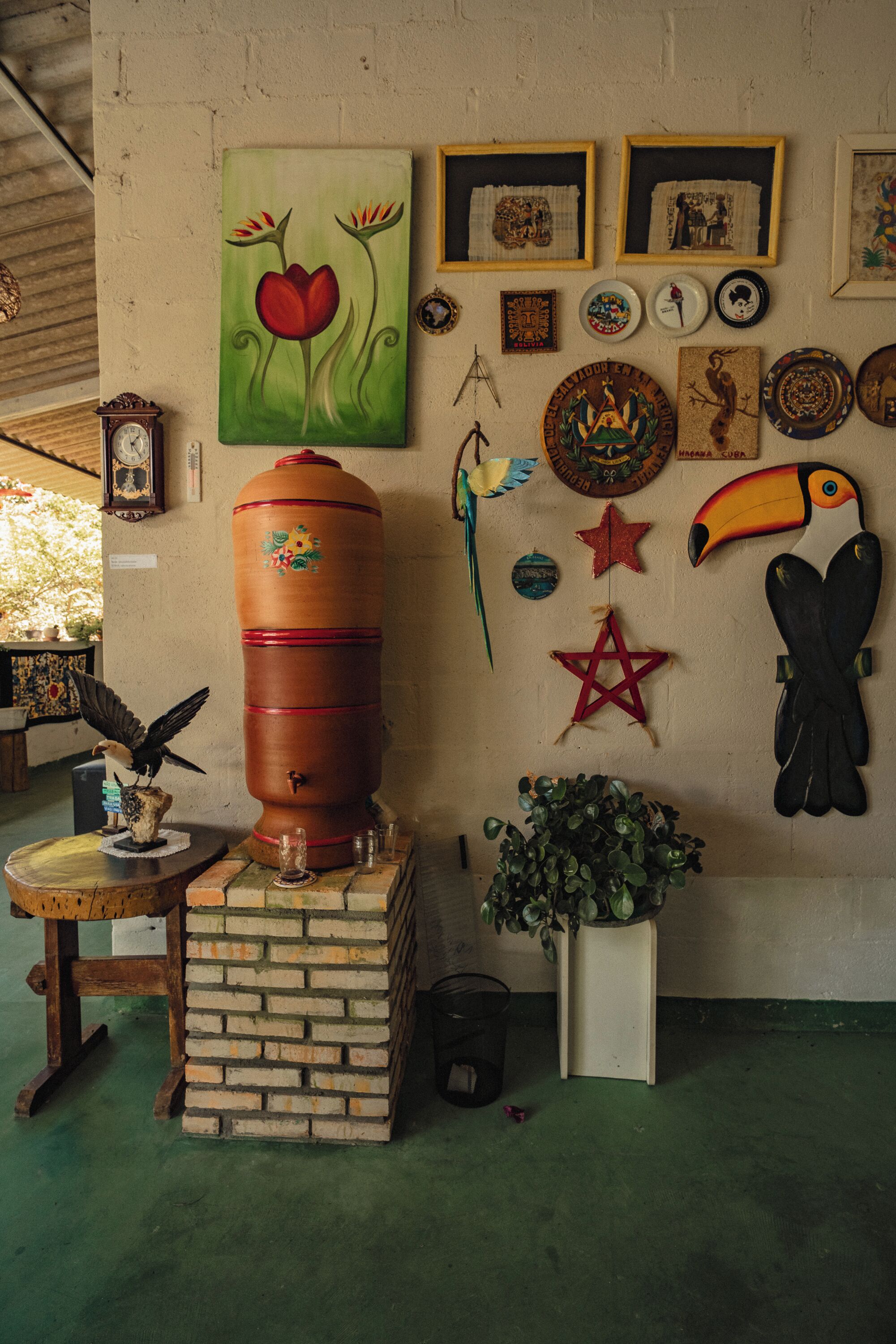A room with a large wooden barrel, a small table with a bird sculpture, a potted plant, and various wall decorations including a star, a toucan, and a painting of flowers.