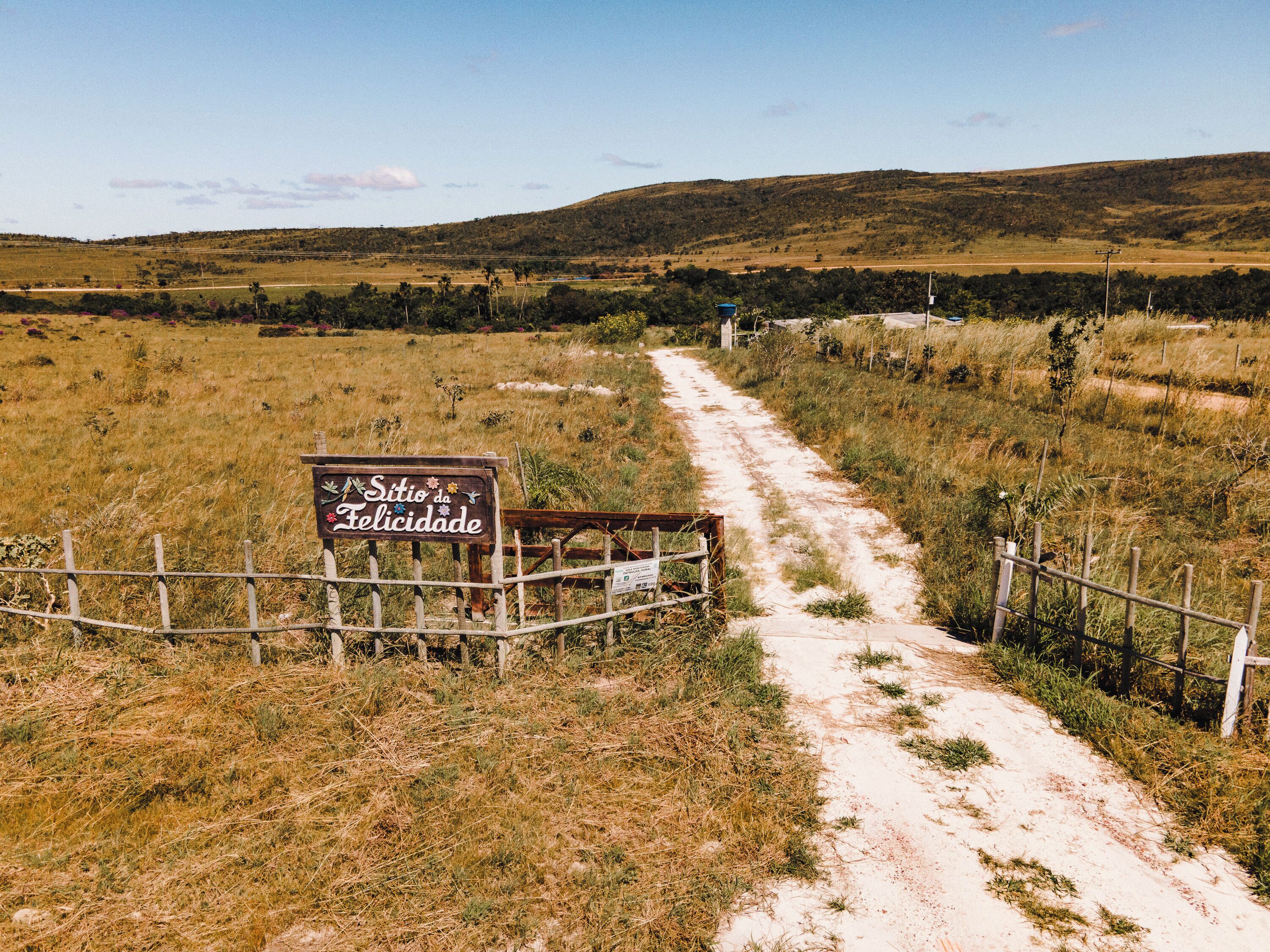 Un sendero de tierra que lleva a un letrero que dice "Sítio da Felicidade", con una cerca de madera y vegetación escasa.