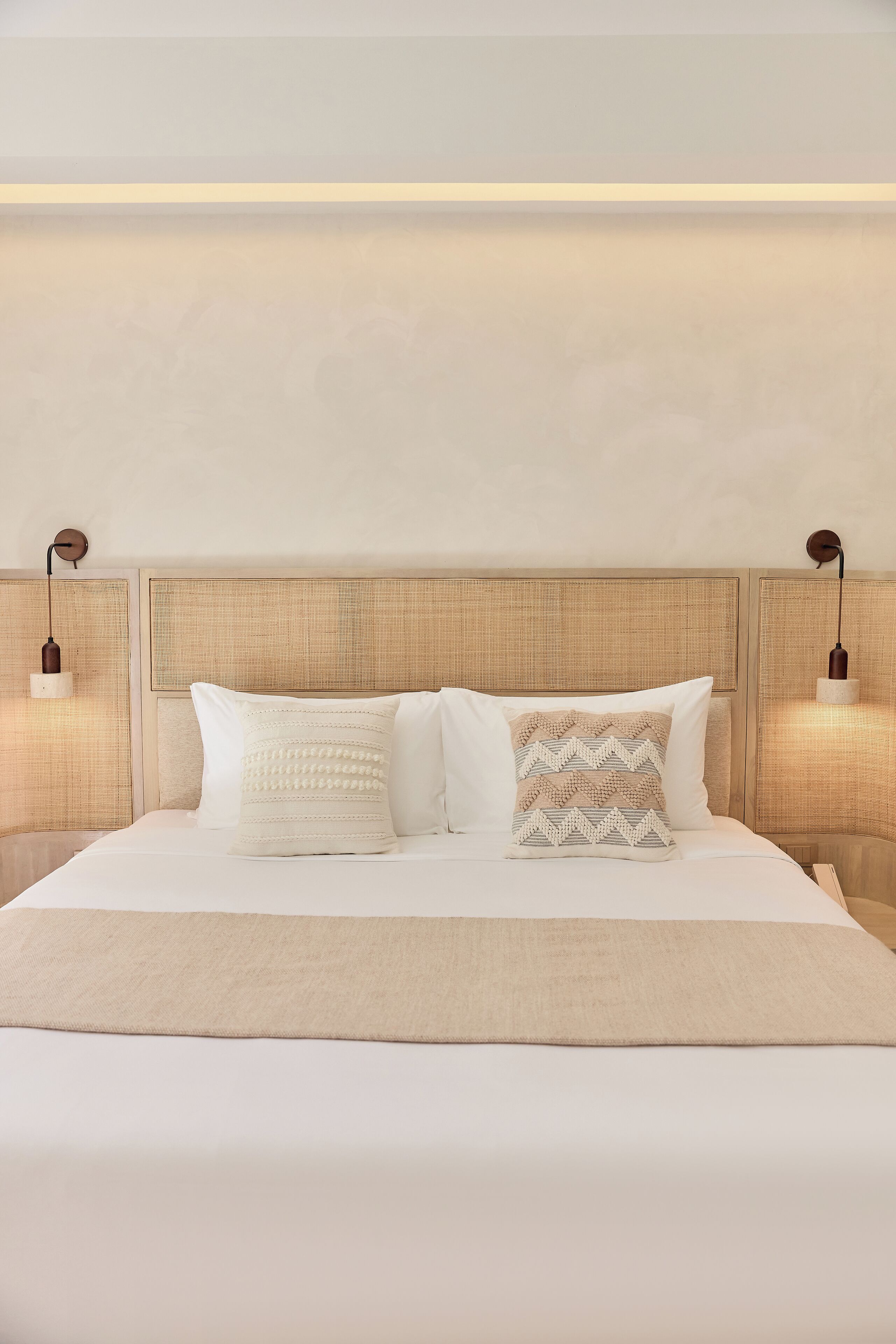 A neatly made bed with a beige blanket and two pillows, a wooden headboard, and two wall-mounted lamps.