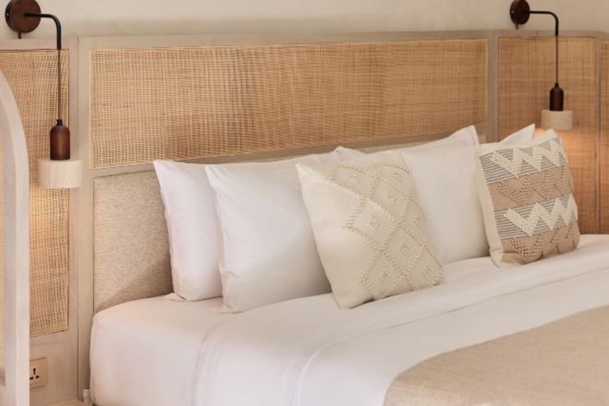 Una cama con ropa de cama blanca, almohadillas decorativas, cabecera de mimbre y apliques de pared.