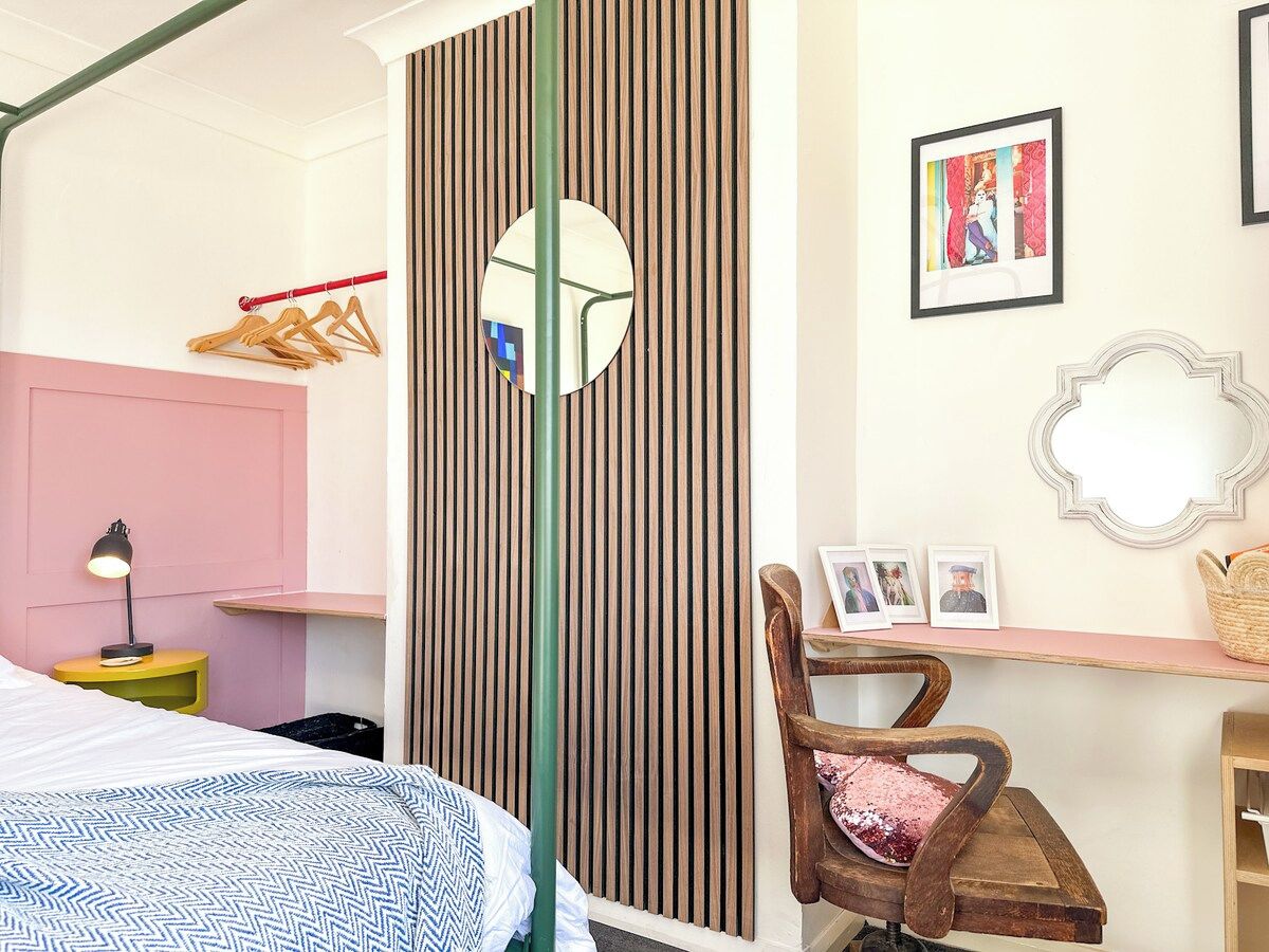 A bedroom with a bed, a bedside table with a lamp, a chair, a mirror, and a wall with vertical wooden slats.