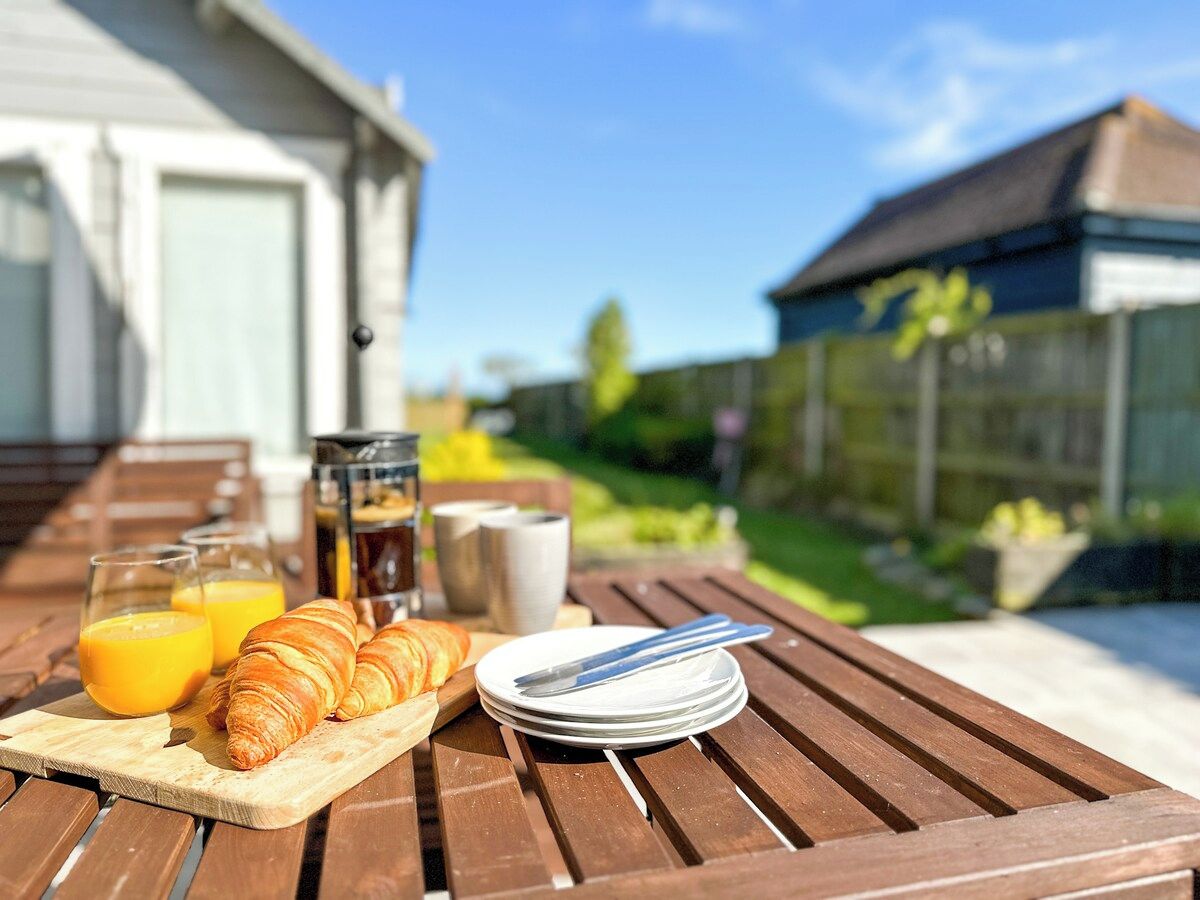 Een houten tafel met ontbijtartikelen zoals croissants, sap en koffie, buiten opgesteld met een huis en groen in de achtergrond.