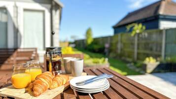 Een houten tafel met ontbijtartikelen zoals croissants, sap en koffie, buiten opgesteld met een huis en groen in de achtergrond.