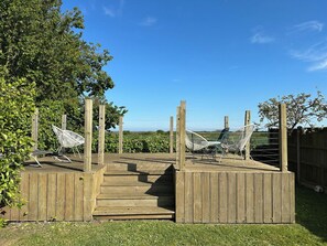 Une terrasse en bois, avec une rampe menant à une plateforme surélevée, entourée d’une clôture en bois et de verdure.