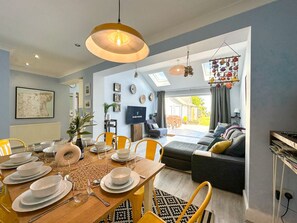 A dining area with a wooden table set for four, a sofa, and a TV in the background. - Modern Family Home near Reculver | Pets Welcome! (Kent)