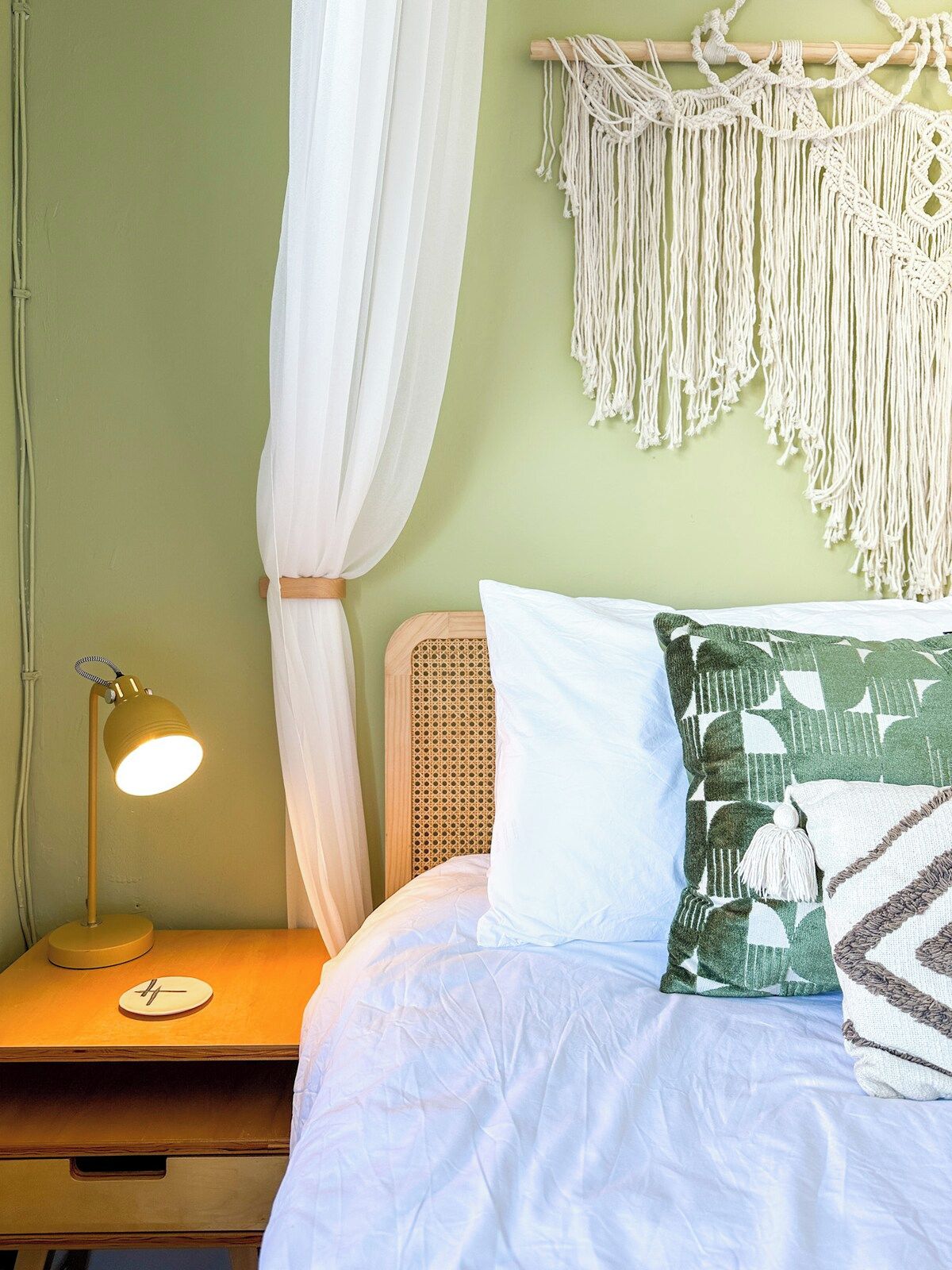 Un lit avec une literie blanche, un coussin à motifs verts, une décoration murale en macramé et une lampe de chevet.