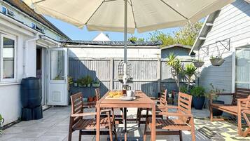 Eine Terrasse im Freien mit Holztisch und -stühlen, einem großen Sonnenschirm und Pflanzkübeln.