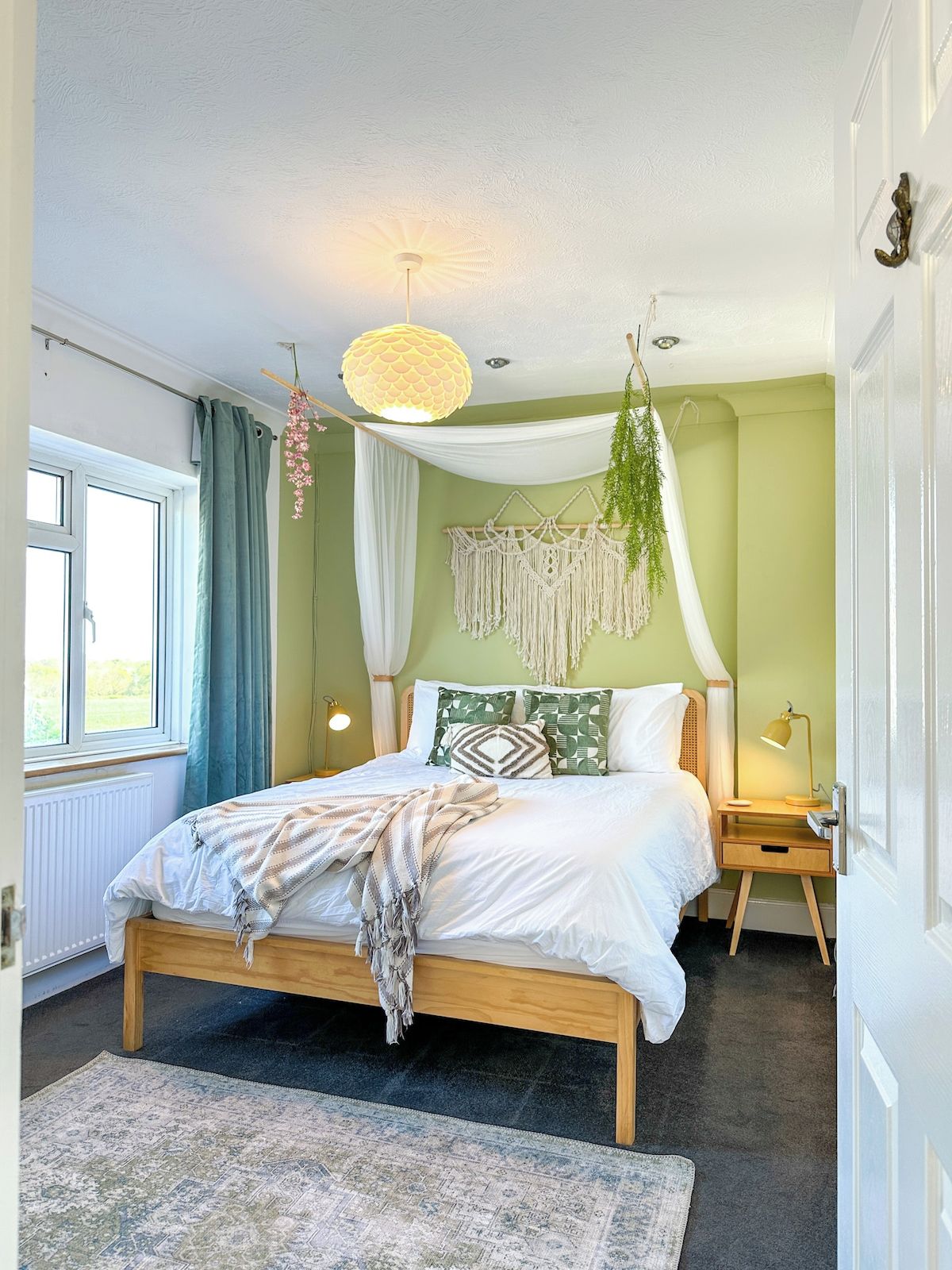 A bedroom with a bed, bedside table, lamp, hanging plants, macrame wall hanging, and a view of the outdoors.