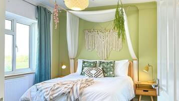 A bedroom with a bed, bedside table, lamp, hanging plants, macrame wall hanging, and a view of the outdoors.