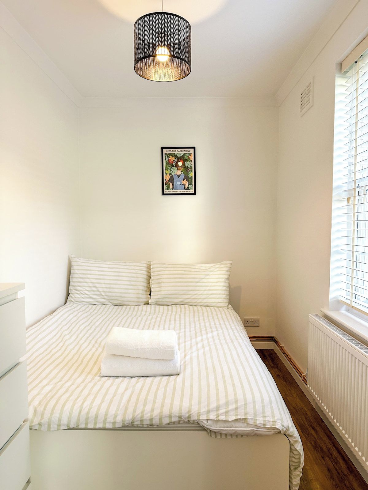 A bedroom with a bed, a hanging light fixture, a framed picture on the wall, and a window with blinds.