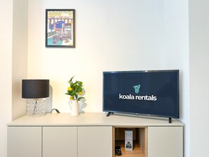 A television displaying 'koala rentals' on a white wall, a lamp, and a potted plant with yellow flowers.
