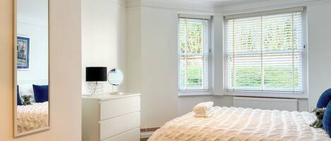 Une chambre à coucher comprenant un lit, une commode, un miroir et deux fenêtres équipées de stores.