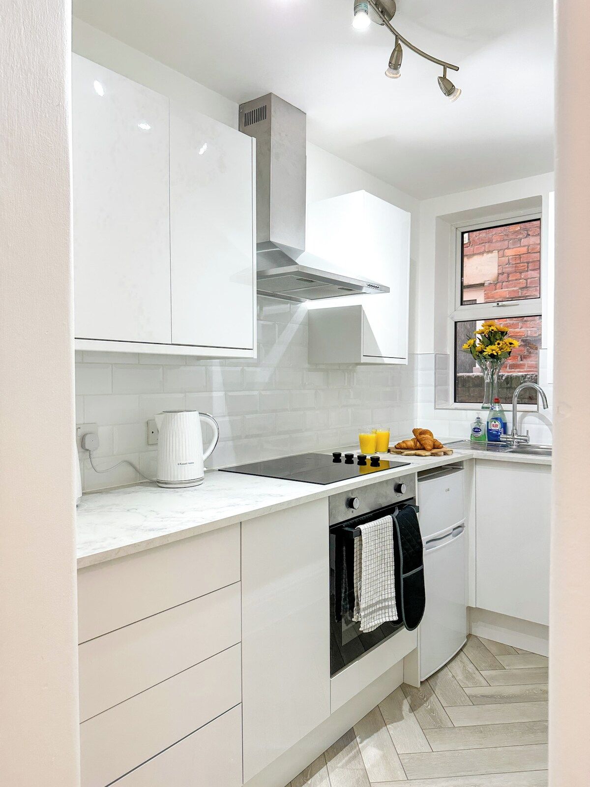 Une cuisine moderne avec des meubles blancs, un four encastré, une bouilloire et une fenêtre donnant sur un mur de briques.