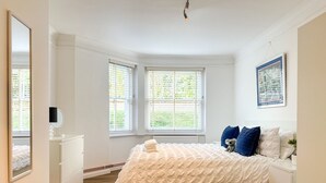Un dormitorio con una cama grande, una mesita de noche, un aparador y dos ventanas con persianas.