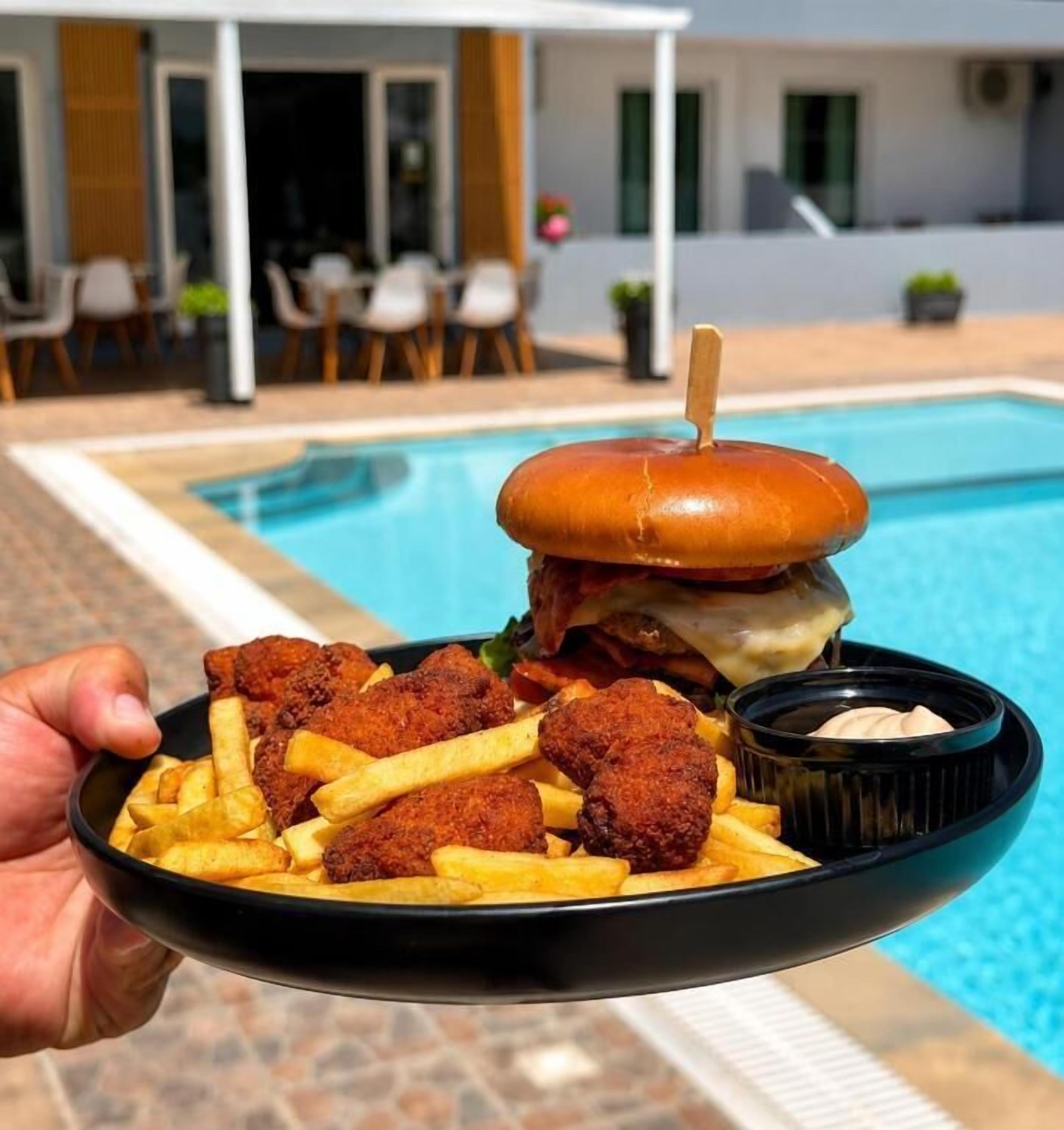 Eine Person hält einen Teller mit einem Burger, Pommes und einem Getränk, im Hintergrund ist ein Pool und Sitzgelegenheiten im Freien zu sehen.
