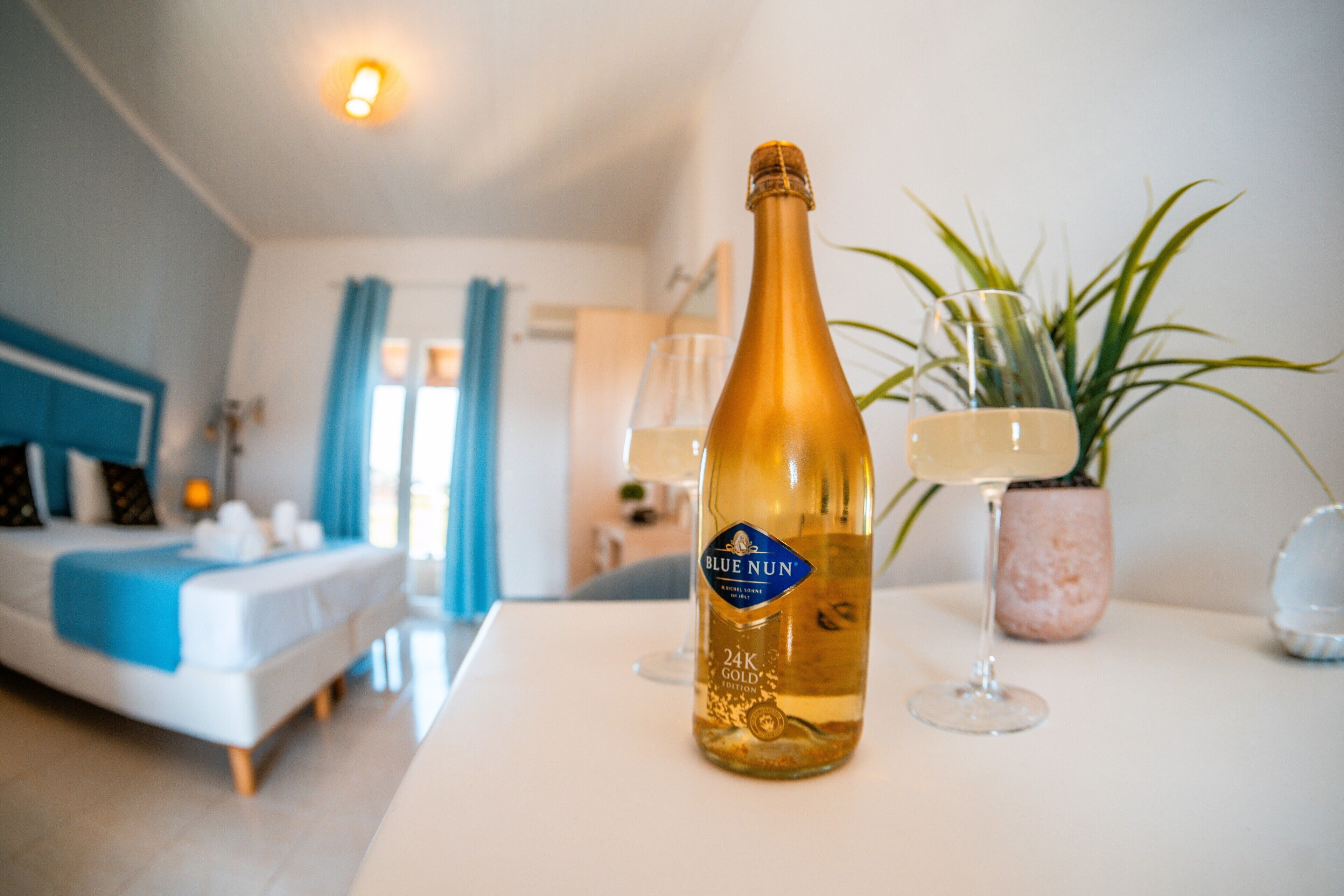 A bottle of Blue Nun champagne, a bed, a bedside table, a potted plant, and a lamp.