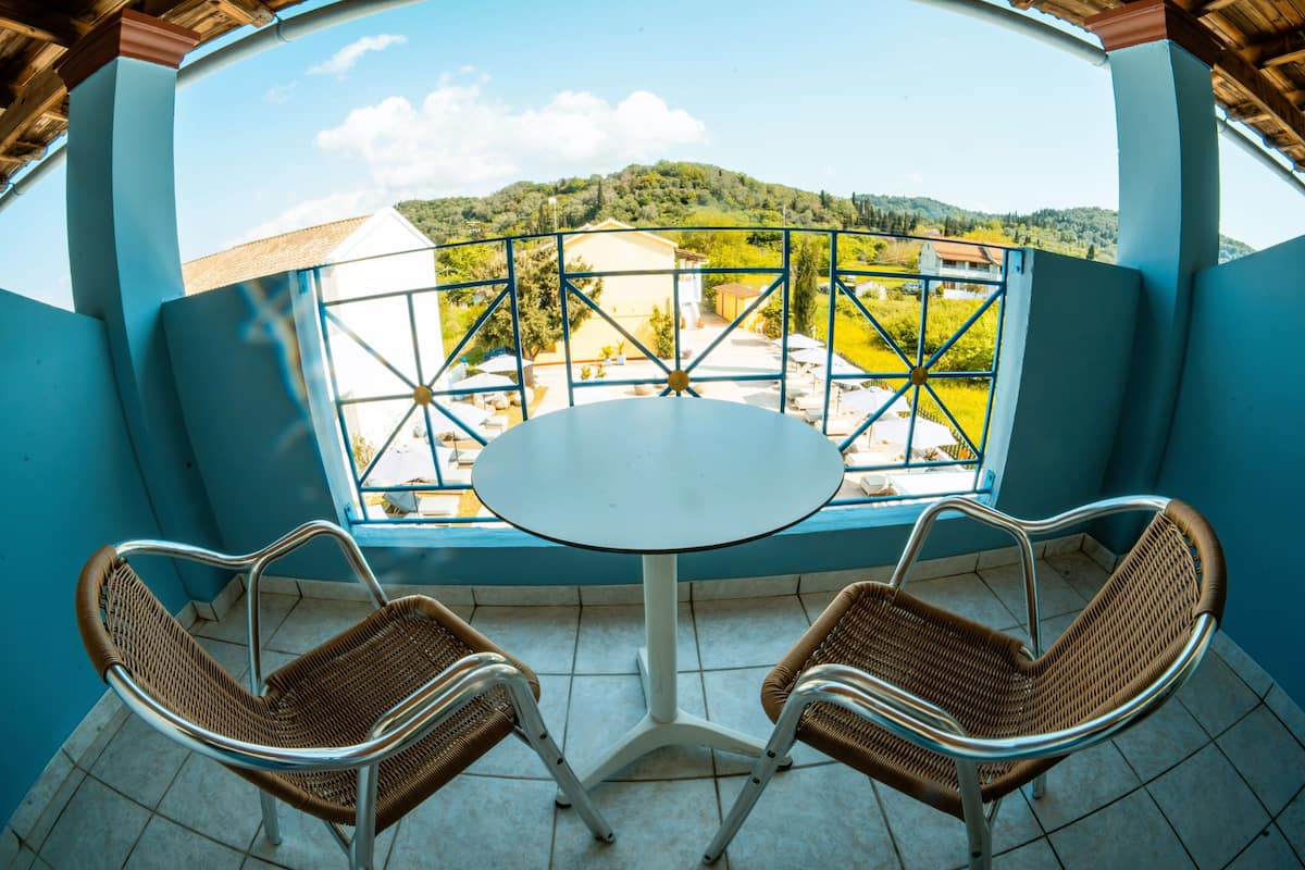 A balcony with a table and two wicker chairs, overlooking a green landscape and buildings.