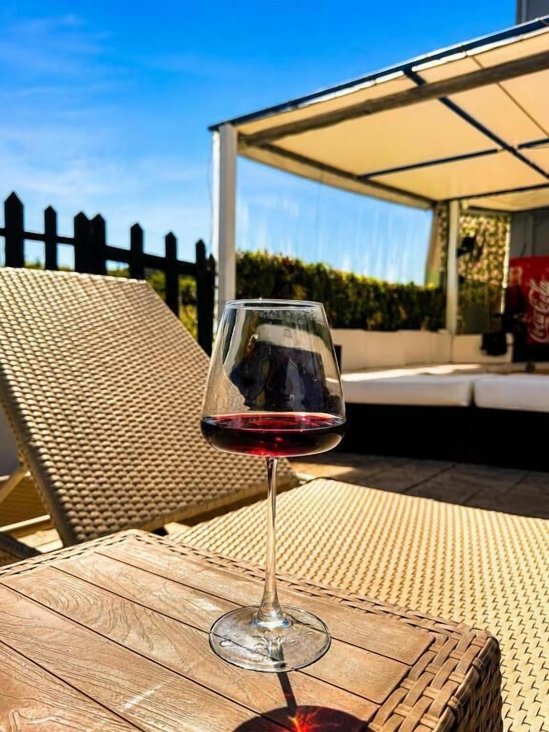 A glass of red wine on a wooden table with a wicker chair and a covered outdoor seating area.