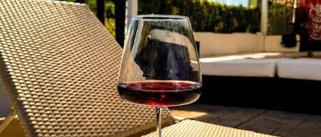 A glass of red wine on a wooden table with a wicker chair and a covered outdoor seating area.