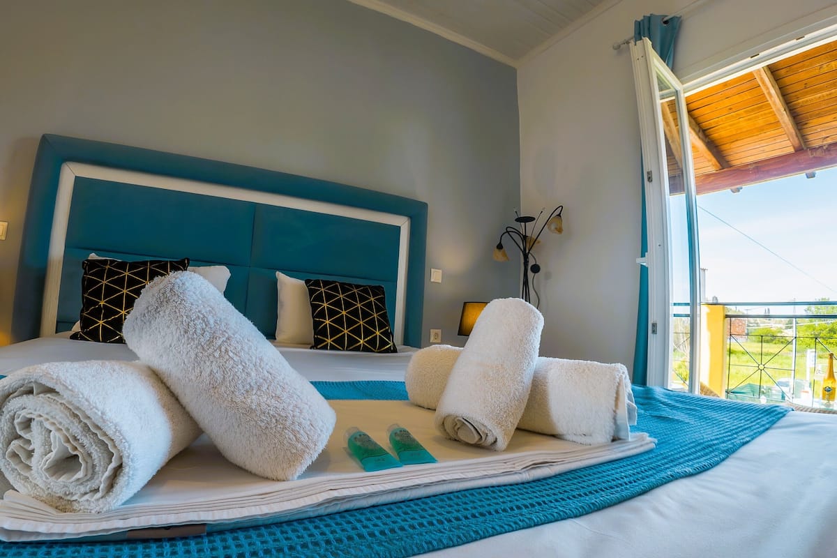 A neatly made bed with towels, a blue headboard, and a view of greenery outside.