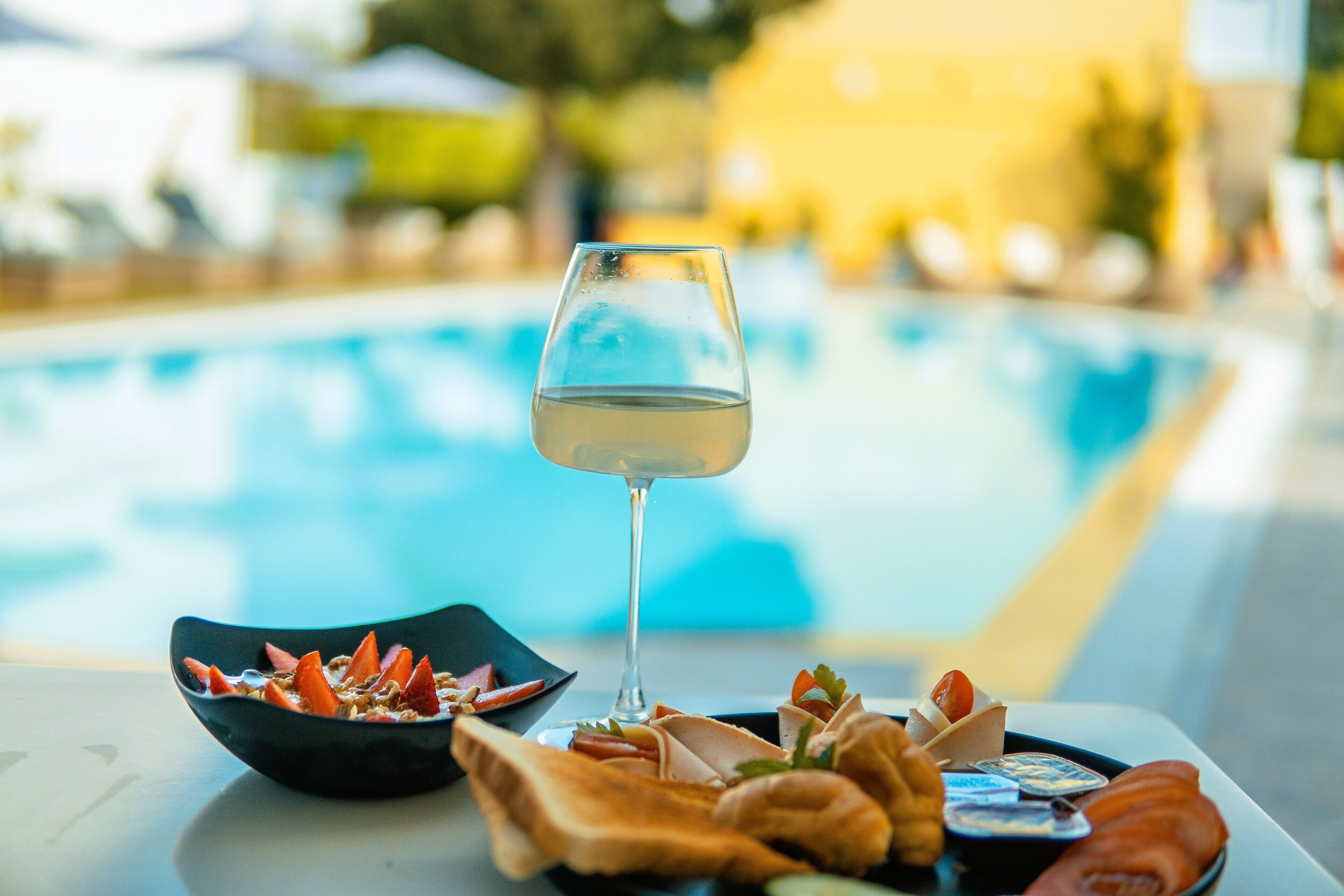 A glass of white wine, a bowl of granola with fruit, and a tray of bread and cheese on a table by a pool.