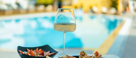 A glass of white wine, a bowl of granola with fruit, and a tray of bread and cheese on a table by a pool.