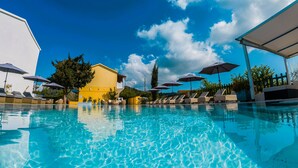 Una piscina con sillas de descanso y sombrillas, un edificio amarillo y un cielo azul despejado.