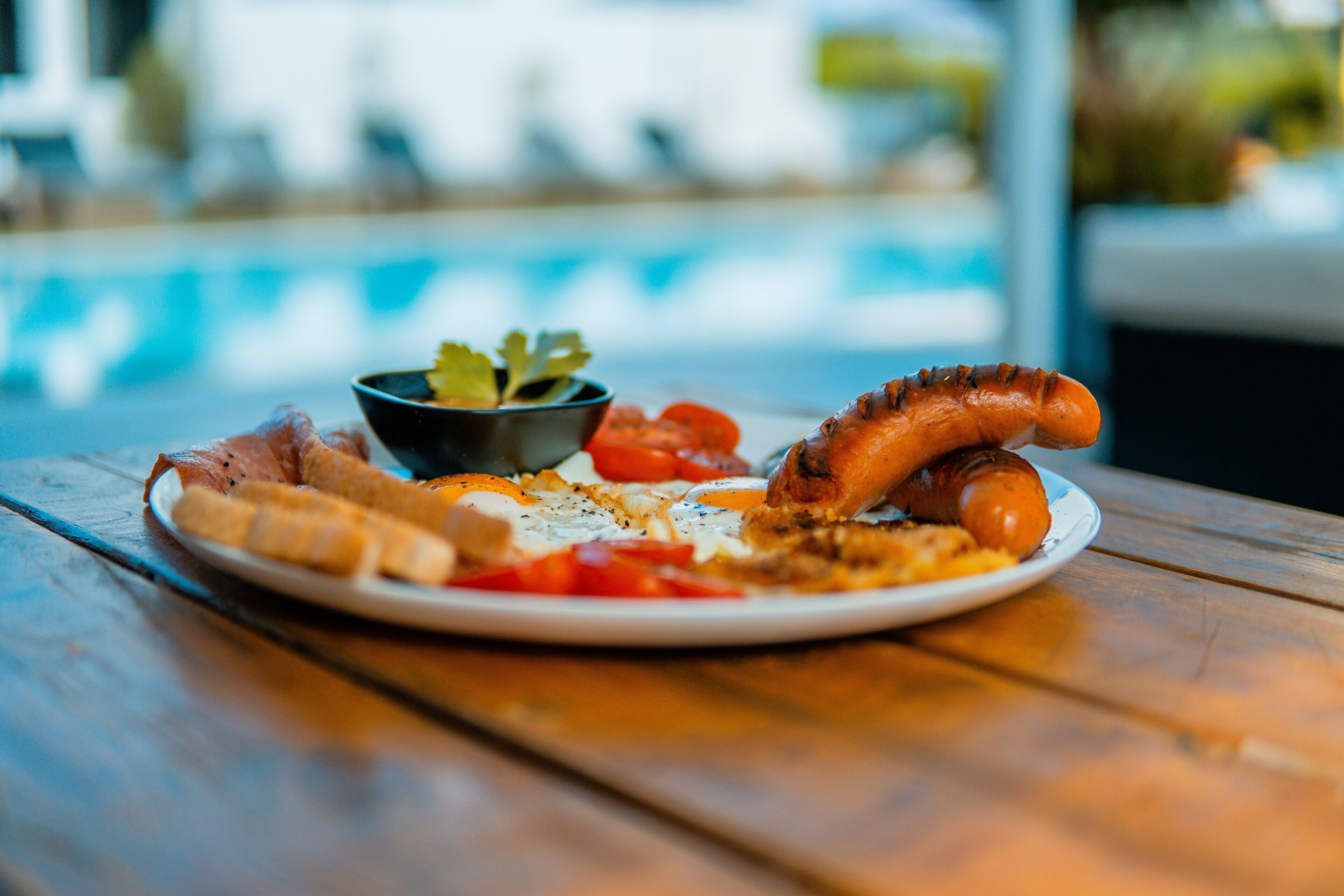 A plate of food with sausage, cheese, and vegetables on a wooden table by a pool.