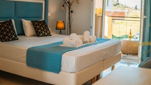 Una cama bien hecha con una manta azul, almohadas blancas y toallas, una lámpara de pie y vistas al exterior.