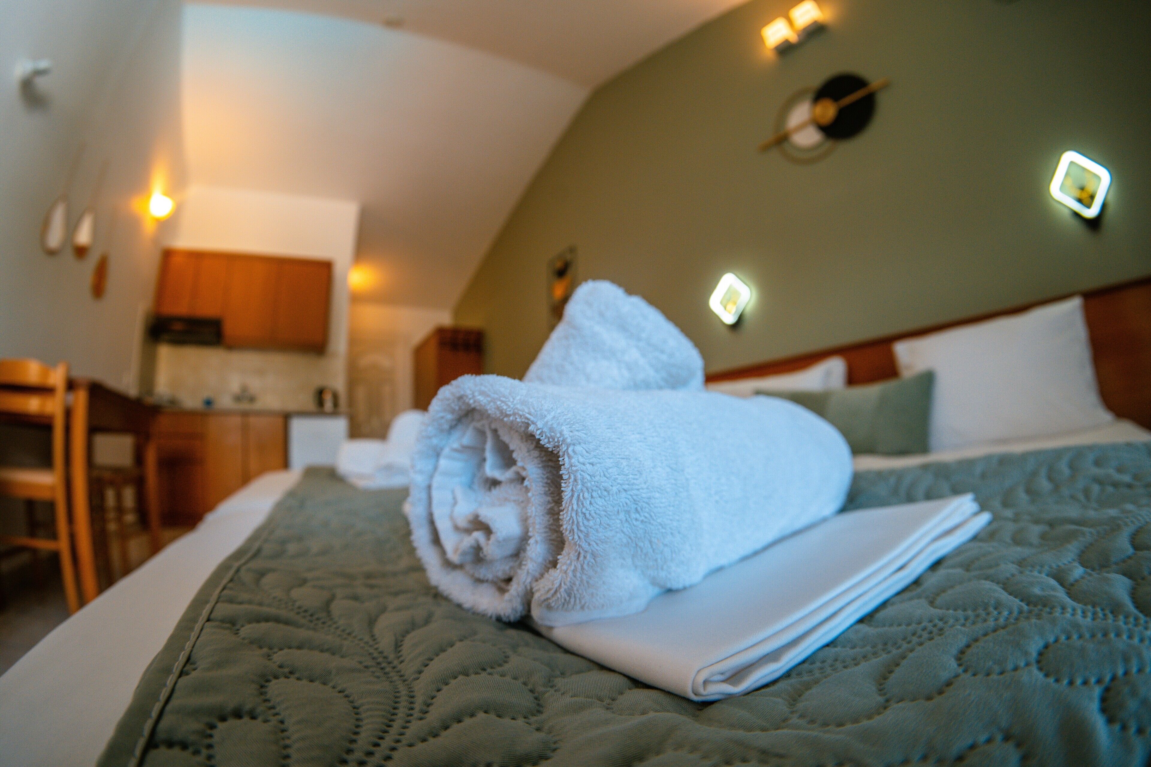 A neatly made bed with a rolled towel and a folded pillow, a wooden headboard, and a kitchen area in the background.